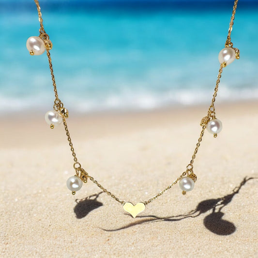 Elegant 18K yellow gold chain necklace with spaced pearls and a heart-shaped pendant, delicately hanging against a soft sandy beach backdrop with a serene blue ocean in the distance.